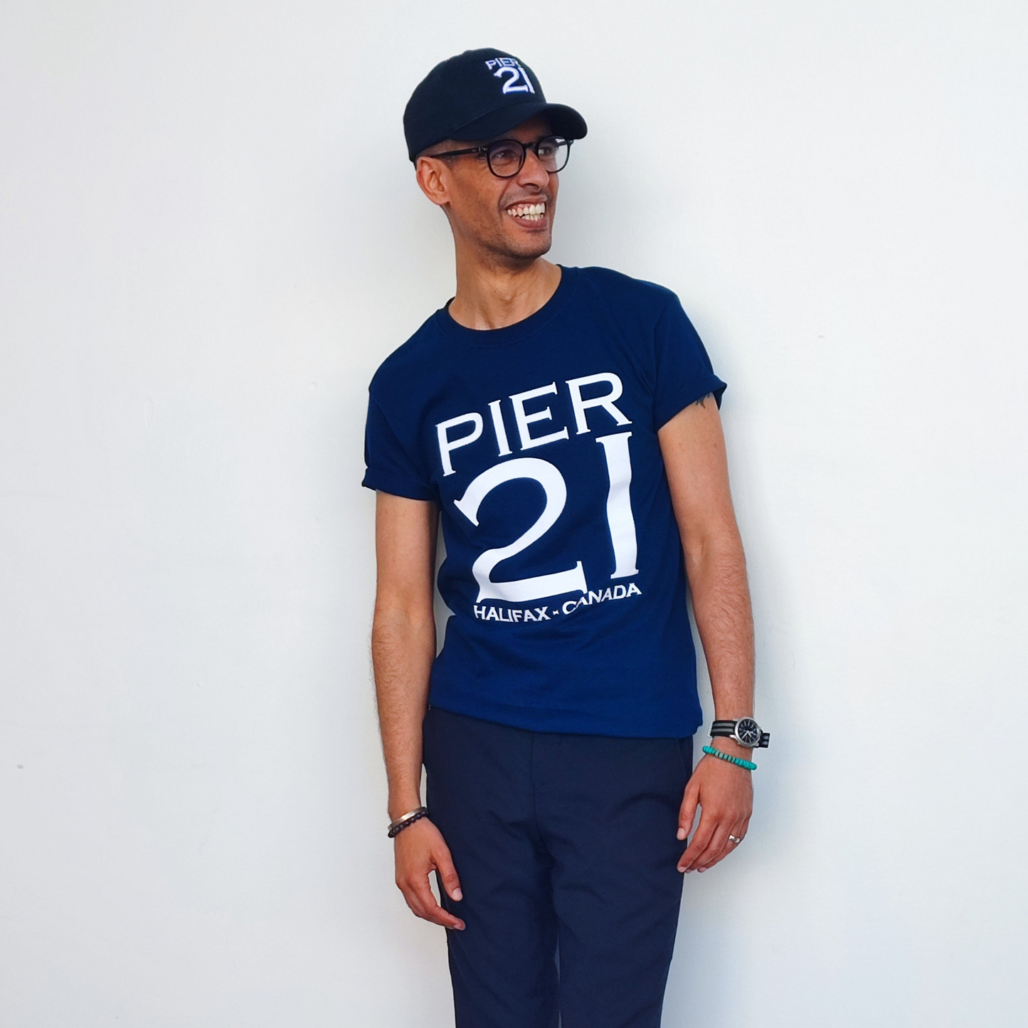 Male model in navy Pier 21 shirt and black Pier 21 hat against a white wall