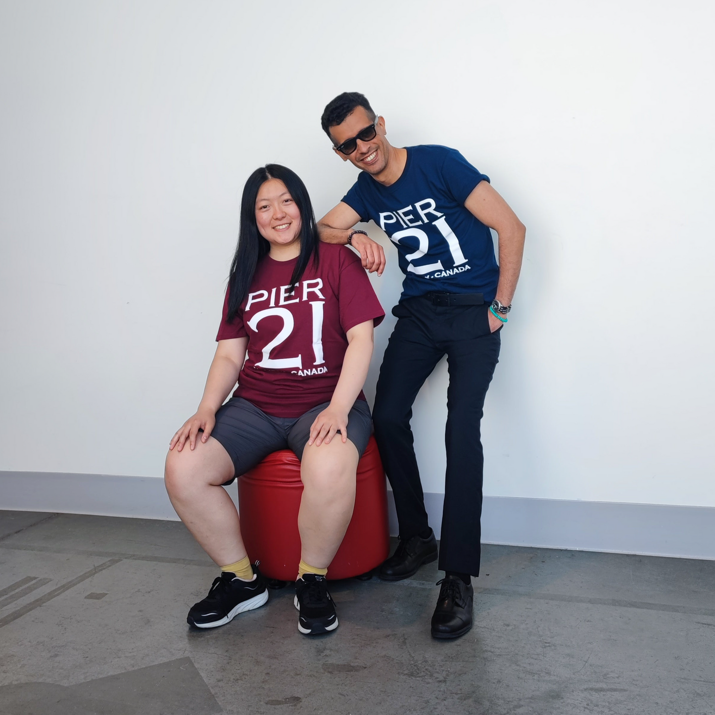 Male model wearing navy Pier 21 shirt leaning on female model wearing Pier 21 burgundy Pier 21 t-shirt who is sitting on red foot stool