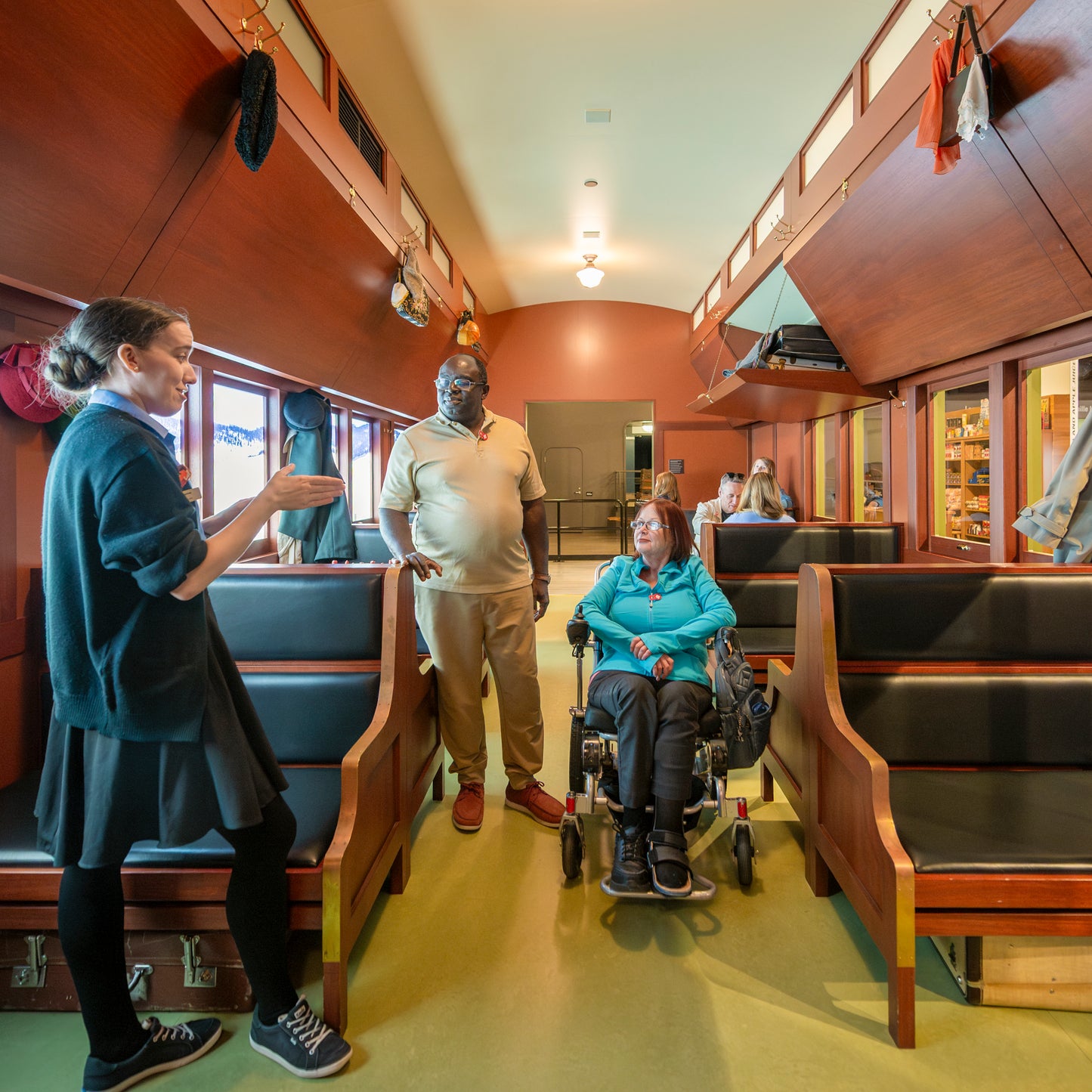 A museum guide speaks with two visitors, one in a wheelchair, inside a historic train car exhibit with wooden benches.
