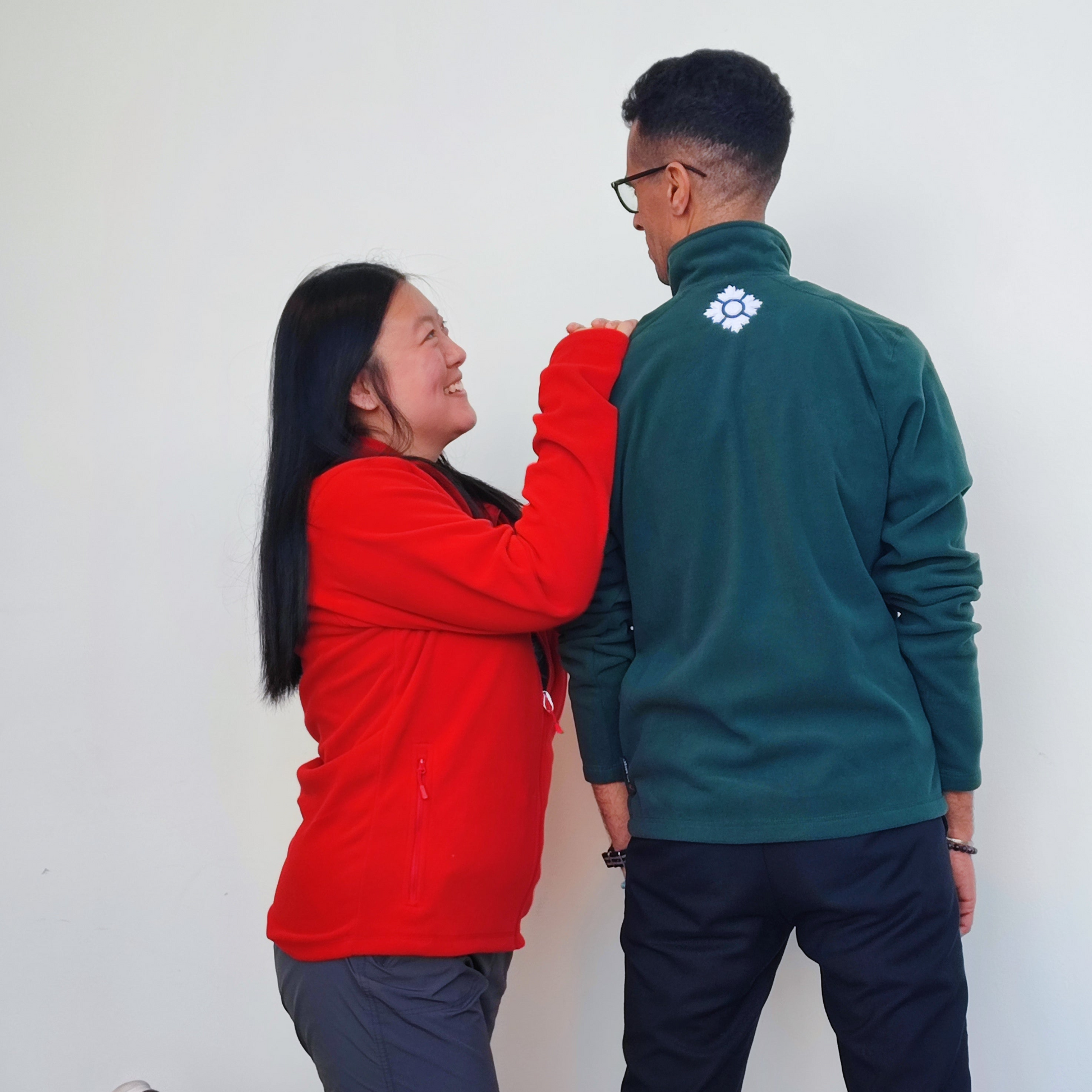 Female model wearing red zip up with arms on male model's shoulder who is facing the wall wearing the green zip up 