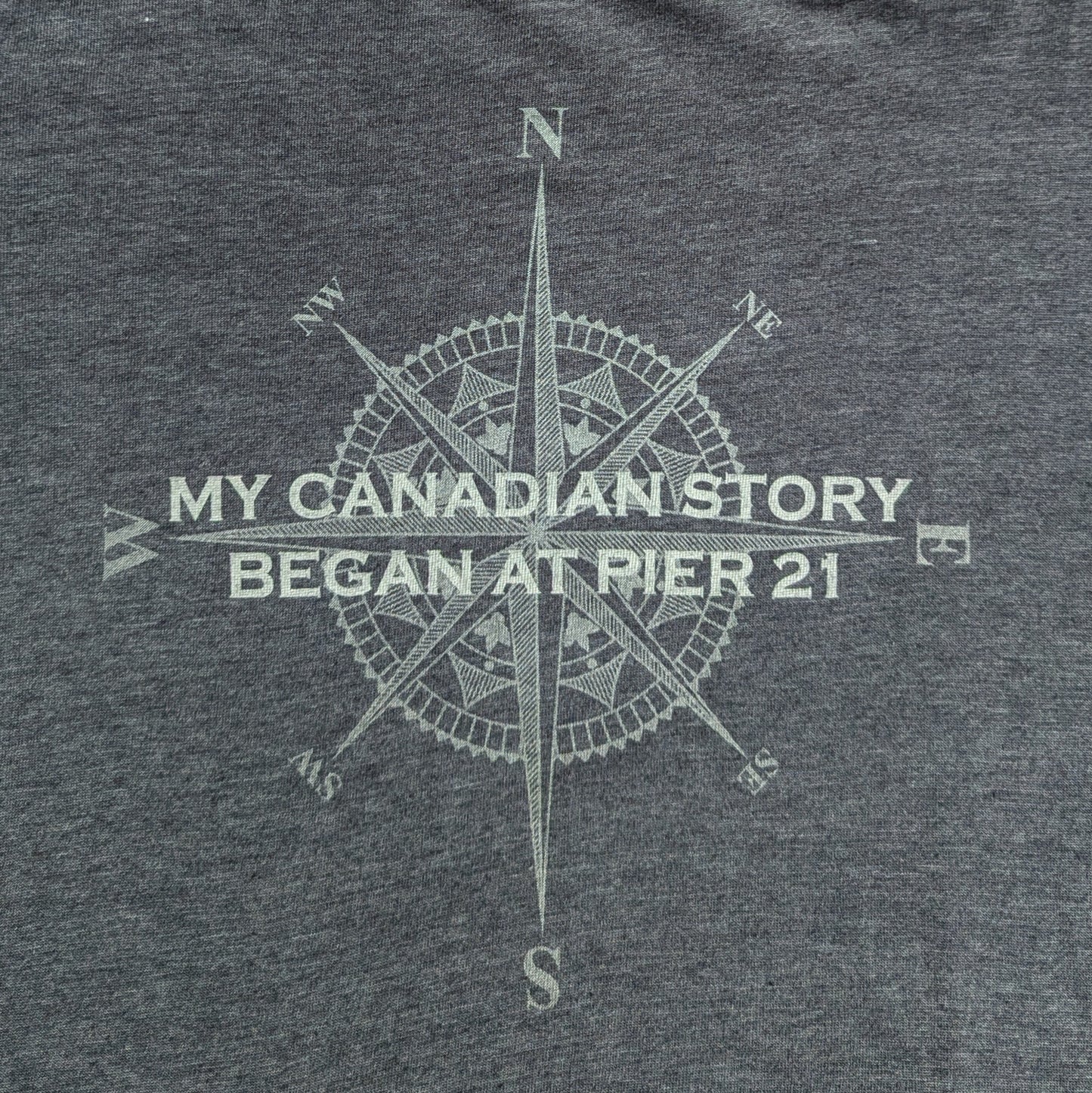 Close up of grey shirt with compass rose graphic with words, My Canadian Story Began at Pier 21 