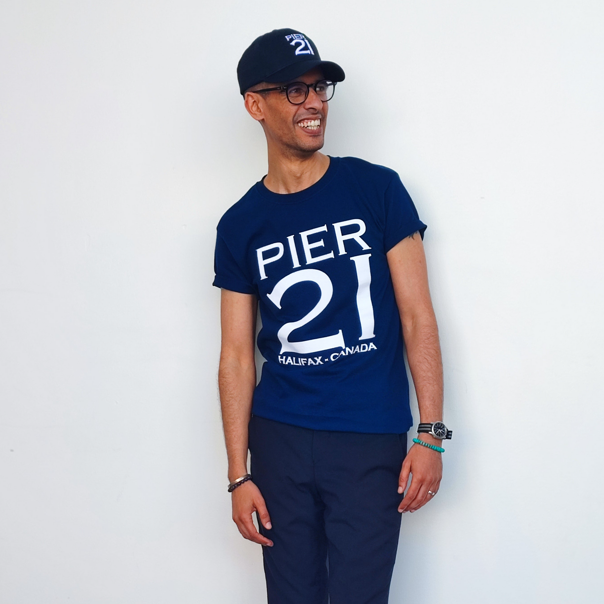 Male model in navy Pier 21 shirt and black Pier 21 hat against a white wall 