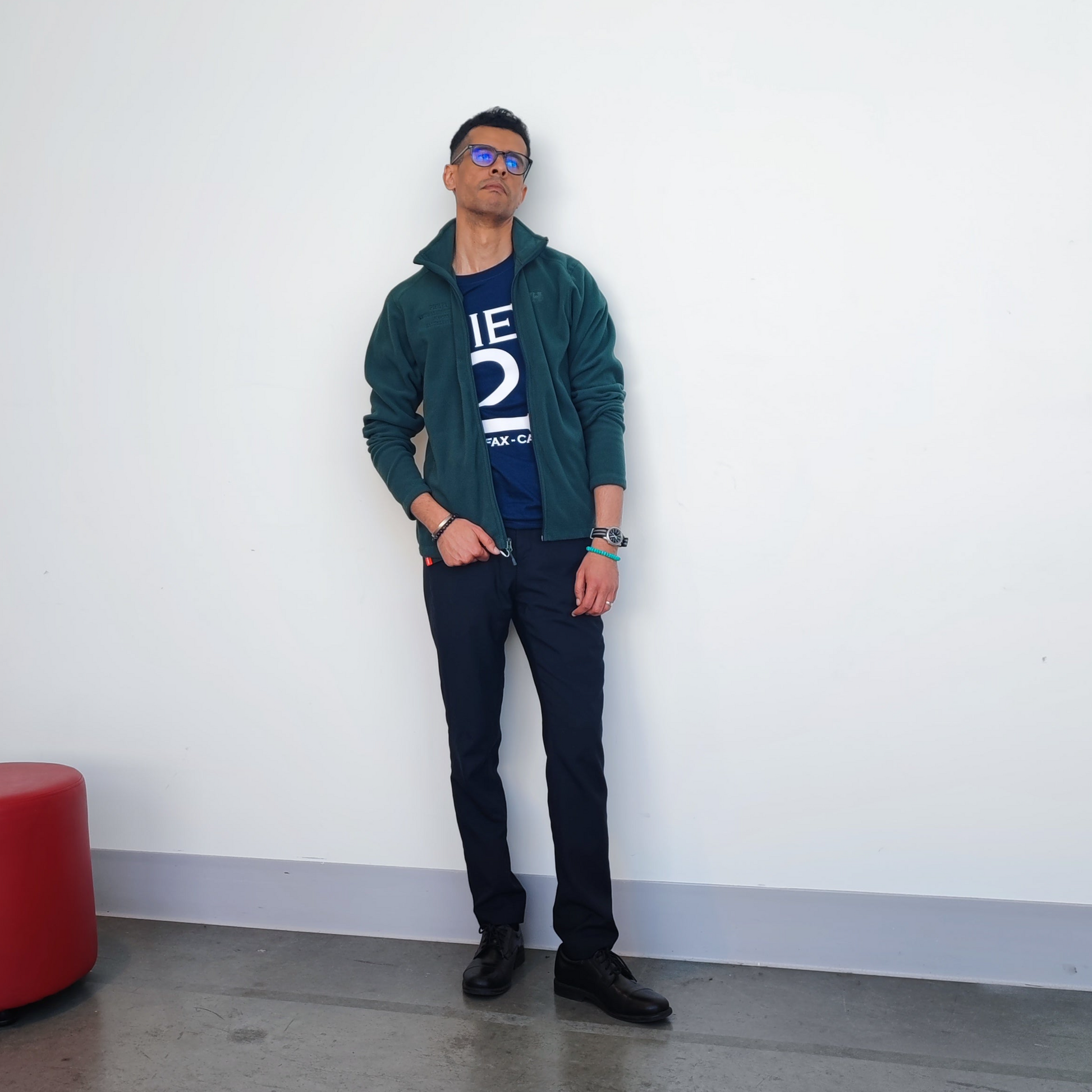 Male model wearing green zip up fleece open over navy Pier 21 t-shirt leaning against a white wall 