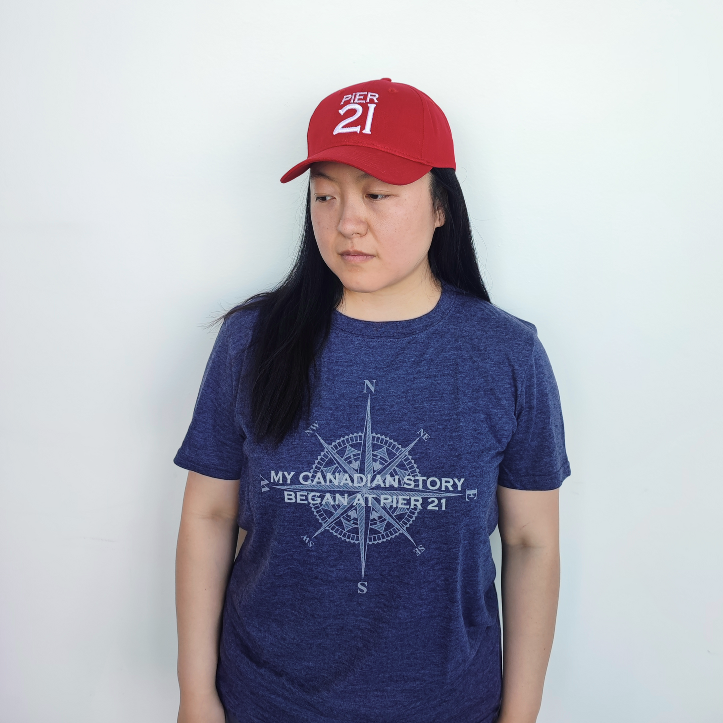 Woman wearing the navy, My Canadian Story Began at Pier 21 t-shirt and red Pier 21 ball cap 