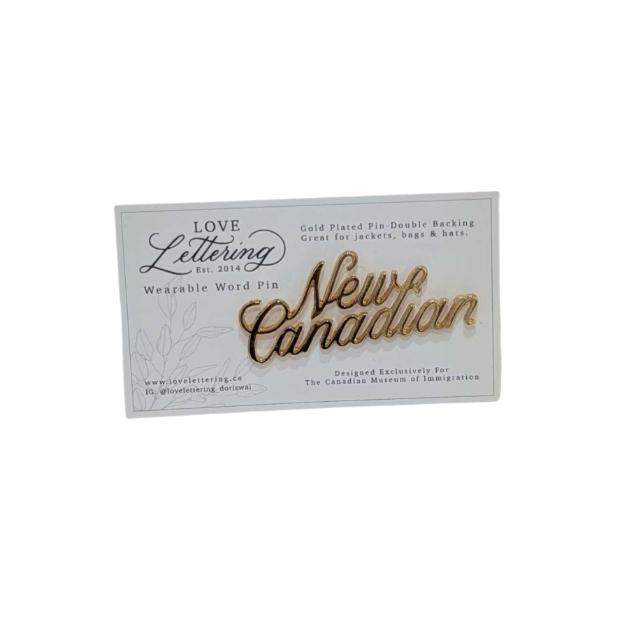 Gold pin saying New Canadian in cursive writing on a white backing card with logo of artisan 