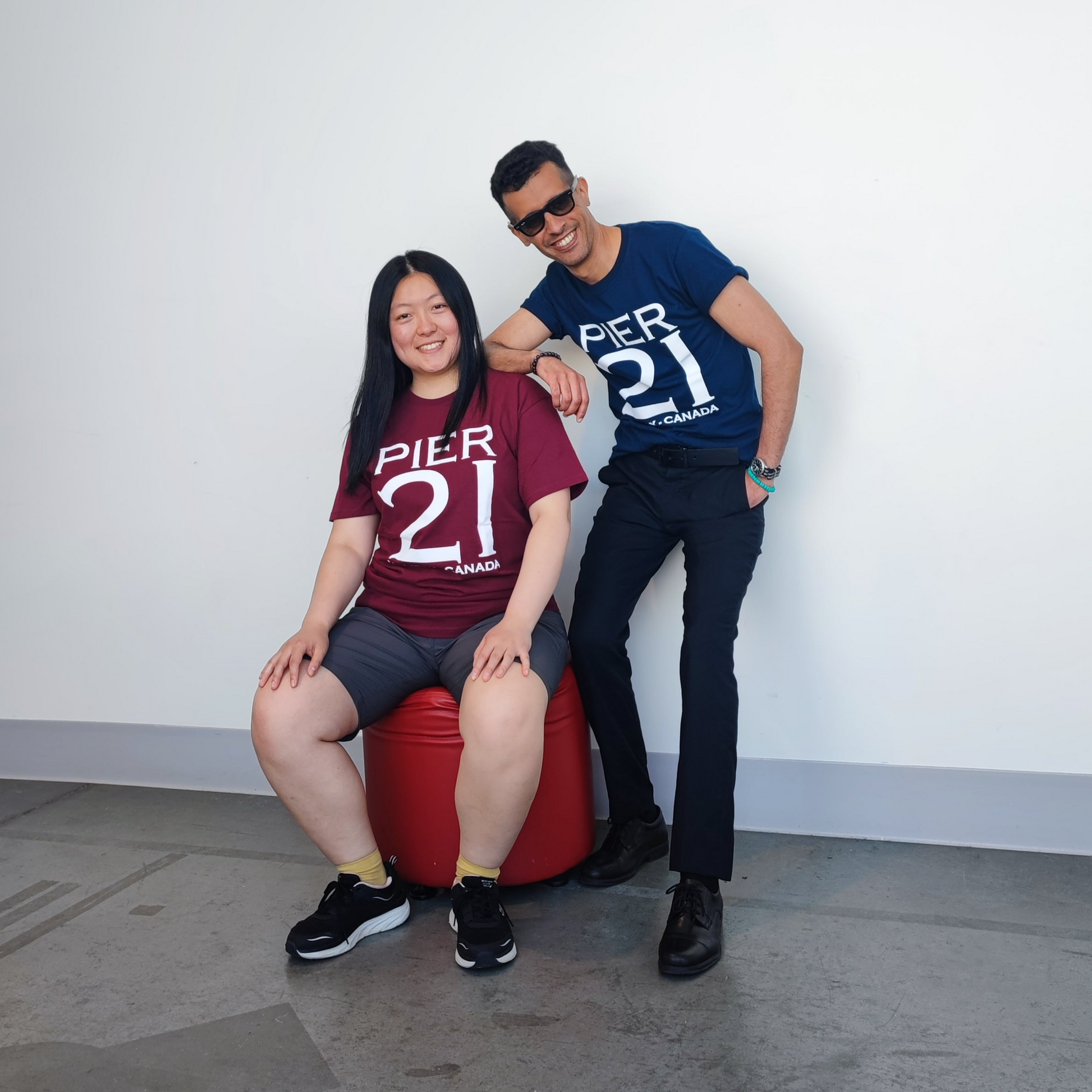 Male model wearing navy Pier 21 shirt leaning on female model wearing Pier 21 burgundy Pier 21 t-shirt who is sitting on red foot stool 