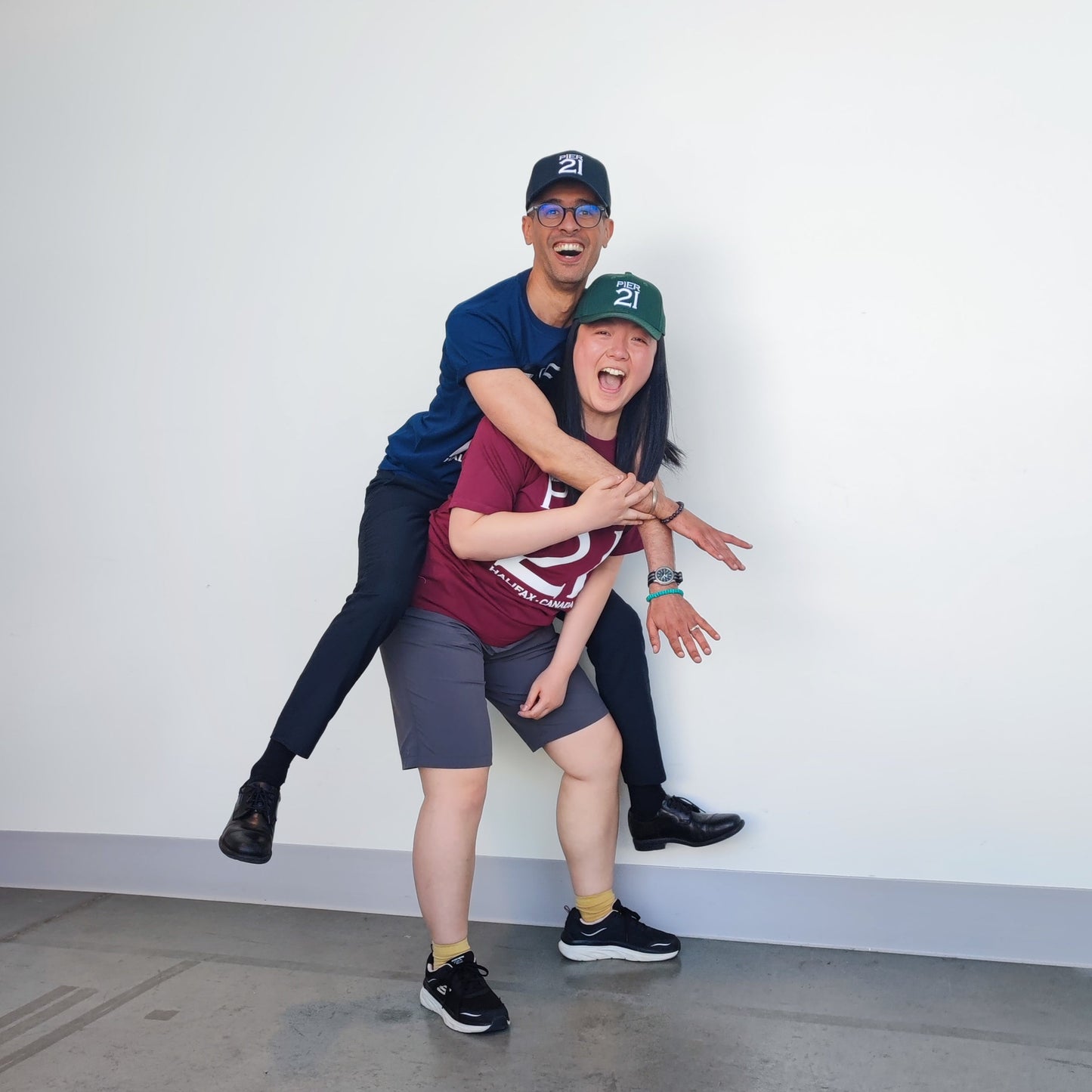 Female model wearing green Pier 21 hat and maroon Pier 21 t-shirt giving a piggy back to a male model wearing a navy Pier 21 hat 