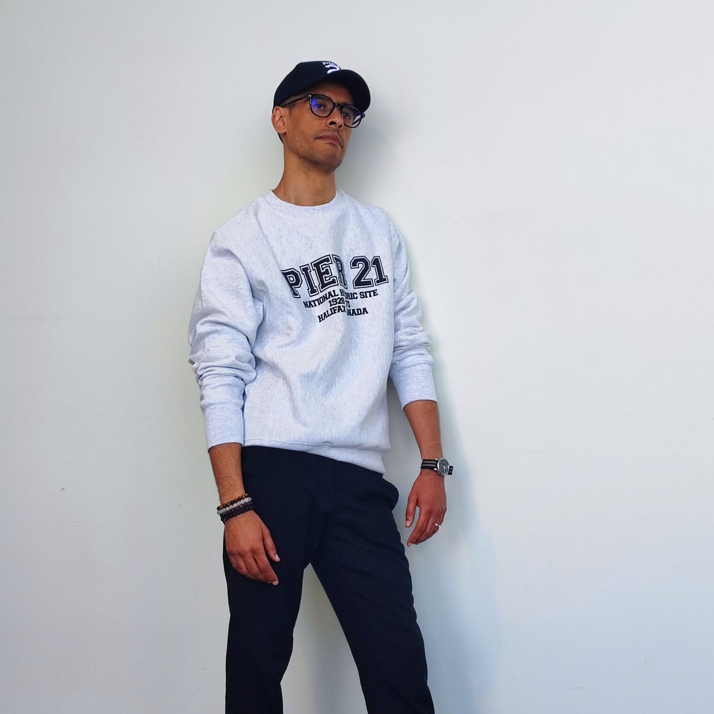 Male model wearing Pier 21 sweatshirt  with sleeves pushed up accompanied by black pants and black Pier 21 cap 