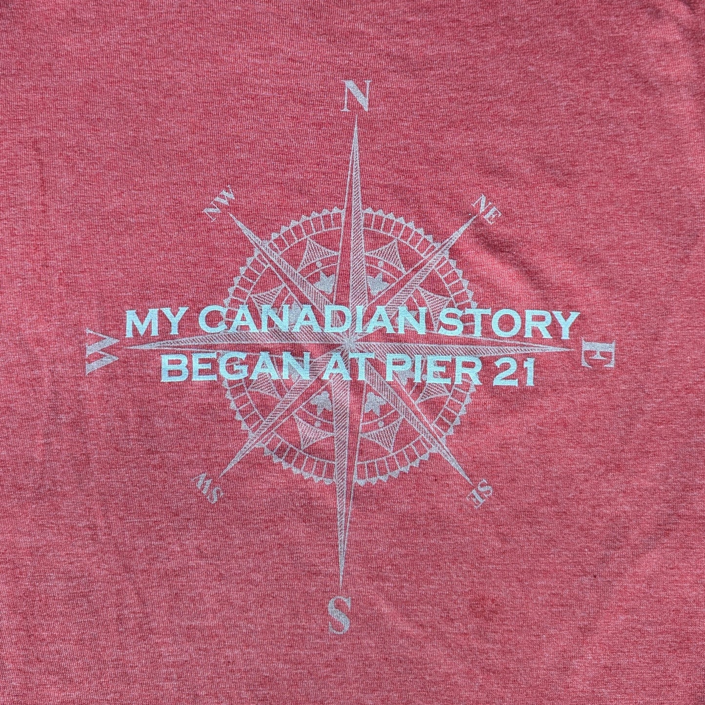 Close up of red shirt with compass rose graphic with words, My Canadian Story Began at Pier 21 