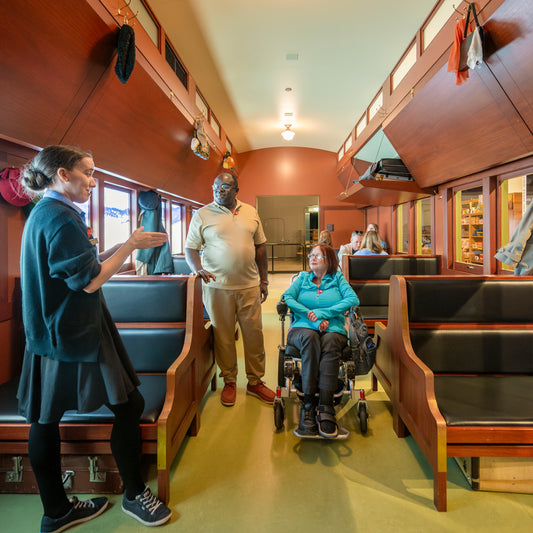 A museum guide speaks with two visitors, one in a wheelchair, inside a historic train car exhibit with wooden benches.