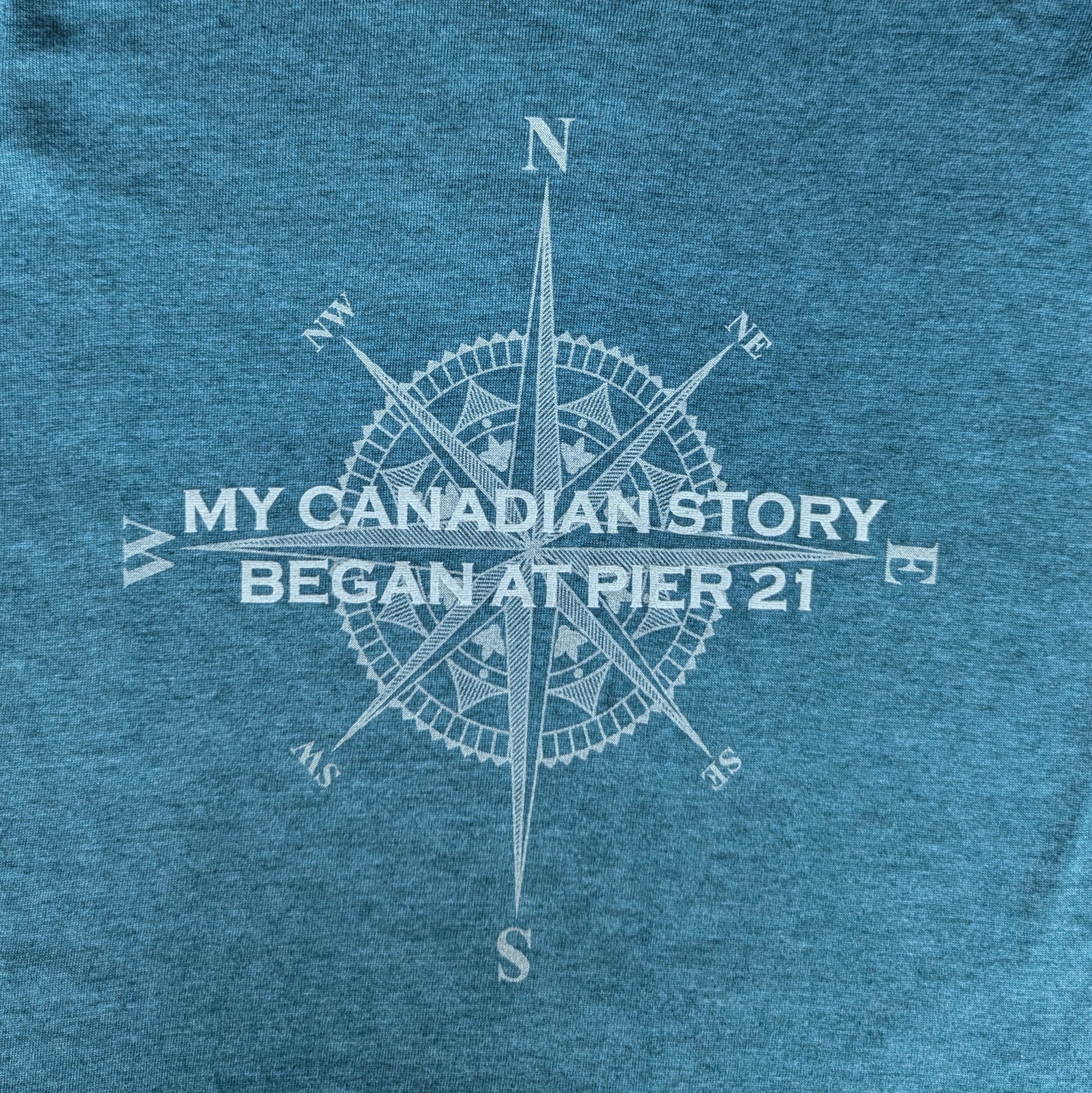 Close up of teal shirt with compass rose graphic with words, My Canadian Story Began at Pier 21 