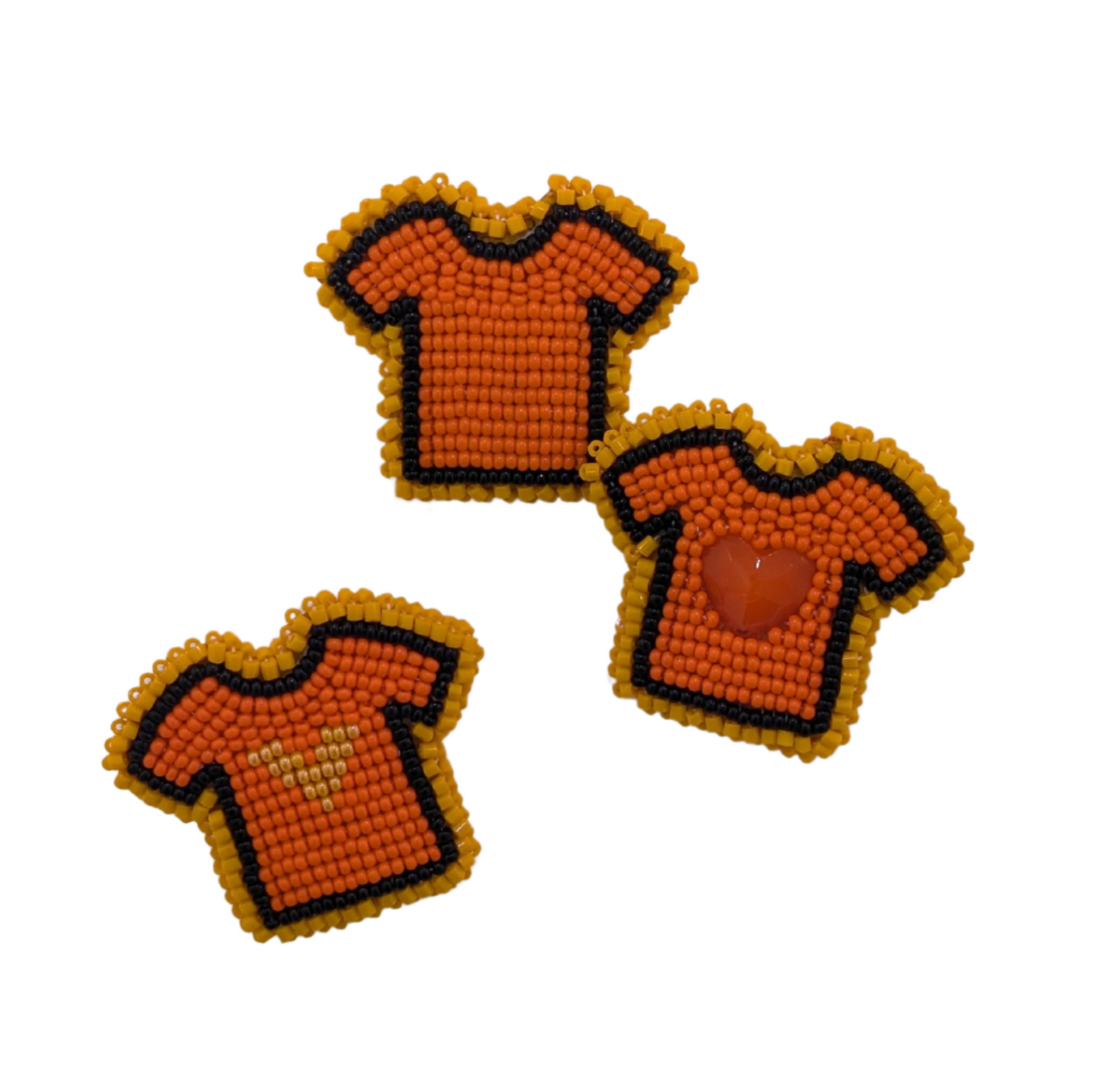 Three orange shirt pins showing the variations in designs such as the top one being plain, the middle pin with a large orange, heart shaped bead in the middle and the lowest showing a lighter orange beaded heart in the center