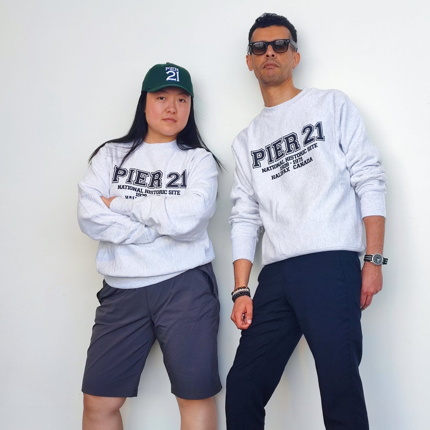 Male and female models wearing Pier 21 sweatshirt leaning against white wall